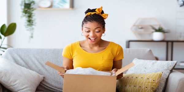 A woman in a yellow shirt opens a box with a surprised look on her face